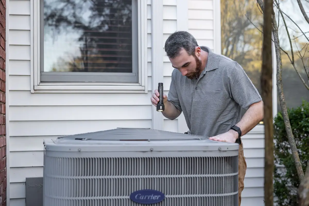A person in a blue jacket and cap kneeling beside an outdoor air-conditioning cabinet.